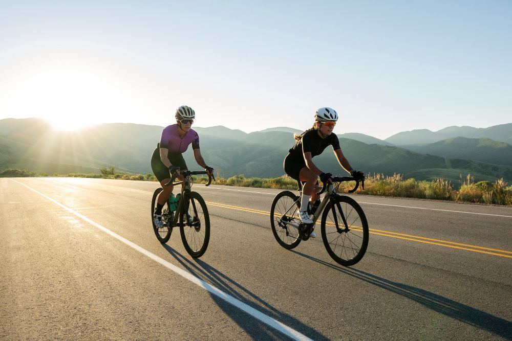 Two road cyclists riding side by side on an open highway at sunrise